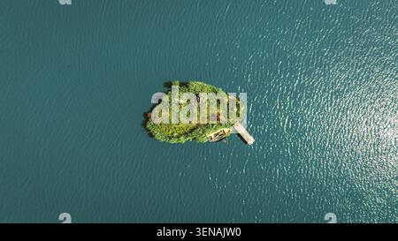 Une île verte isolée couverte de végétation et de fleurs, avec une jetée en bois et un petit bateau, entouré d'une eau bleue tranquille Banque D'Images