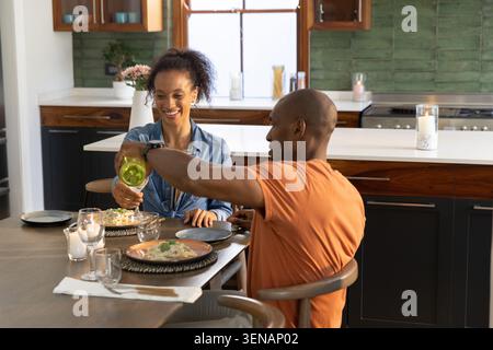 Couple à la table de la cuisine, homme versant de la bouteille de vin vert comme femme tenant l'assiette de pâtes souriant Banque D'Images