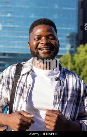 Homme adulte africain debout souriant à l'extérieur de l'immeuble de bureaux en verre portant chemise à carreaux et sac à dos Banque D'Images