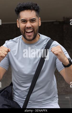 Homme levant les poings et criant sous surplomb, portant des vêtements de gymnastique gris clair, sangle noire, sac, montre Banque D'Images