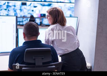 Vue arrière d'une surveillante se tenant debout au-dessus de l'épaule d'un agent de sécurité masculin, le guidant à travers les données sur les écrans d'ordinateur dans une salle de surveillance. Banque D'Images