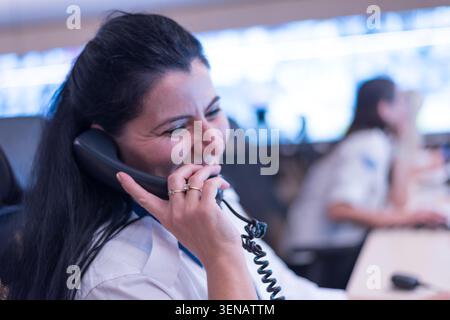Vue de profil d'une joyeuse agente de sécurité aux cheveux foncés souriant tout en parlant sur un combiné téléphonique filaire dans un coffret de salle de contrôle floue Banque D'Images