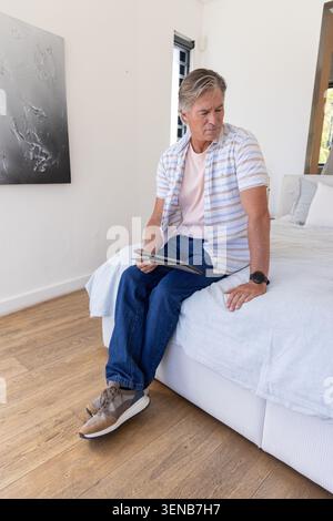 Homme mûr assis sur le bord du lit tenant la tablette, portant une chemise rayée, un Jean bleu, des baskets tan Banque D'Images