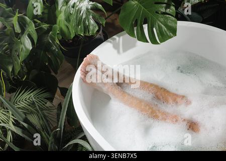 Un spa serein avec des jambes magnifiquement immergées dans une baignoire remplie de bulles, entouré de plantes vertes luxuriantes, parfait pour le bien-être et la relaxation Banque D'Images