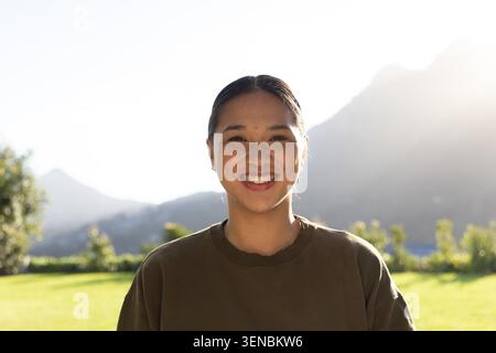 Femme asiatique mi-adulte souriant sur la pelouse avec des arbustes et des montagnes, ensoleillée, portant Crewneck olive Banque D'Images