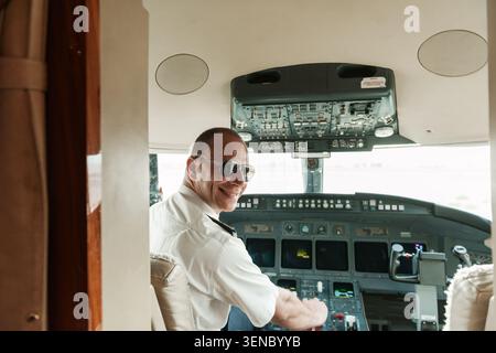 Pilote de ligne confiant en uniforme blanc et lunettes de soleil aviateur souriant sur son épaule aux commandes du cockpit d'un poste de pilotage d'avion idéal pour Banque D'Images