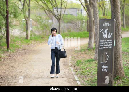 Femme marche le long du chemin du parc, parlant au téléphone. Elle tient une tasse et un sac à main, habillée de façon décontractée. Les arbres bordent le chemin, les feuilles vertes tendres au-dessus. Filtre solaire Banque D'Images