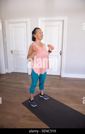 Femme senior soulevant des haltères de corail sur tapis à la maison, portant un haut de corail, leggings sarcelle, smartwatch Banque D'Images