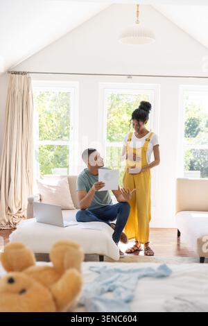 Couple afro-américain tenant l'échographie, souriant au partenaire dans une combinaison jaune dans le salon Banque D'Images