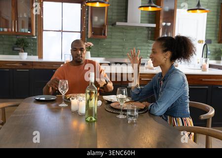 Couple diversifié assis à la table à manger en bois dans la cuisine, coupant la nourriture et parlant sur le vin Banque D'Images
