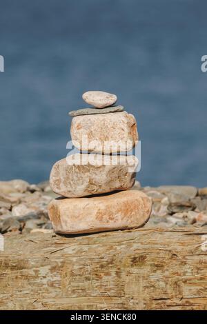 Les pierres empilées s'équilibrent gracieusement sur le bois altéré. Le calme côtier résonne dans chaque surface lisse et arrondie. L'eau bleue s'estompe doucement derrière l'image fixe Banque D'Images