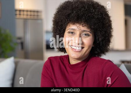 Femme afro-américaine assise sur un canapé gris avec un frigo flou derrière, portant un dessus marron, souriant Banque D'Images