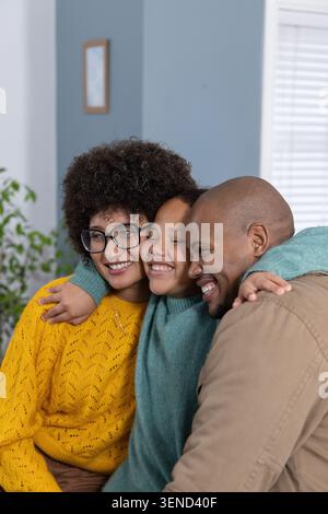 Famille diversifiée embrassant sur le canapé dans le salon, enfant en pull sarcelle, mère portant un tricot jaune Banque D'Images
