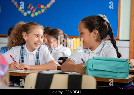 Deux filles joyeuses de l'école primaire assises à leur bureau, souriantes et parlant pendant une leçon de classe. Banque D'Images