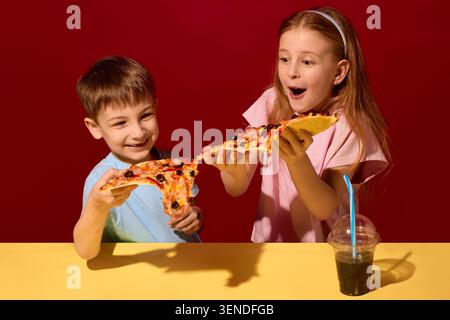 Deux enfants étirant du fromage fondu entre des tranches de pizza sur fond de studio rouge. Banque D'Images