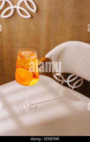 Une boisson avec de la glace et des tranches d'orange se trouve sur une table. Le fond montre un mur chaud et une chaise. La lumière projette une ombre sur le verre, créant une atmosphère détendue. Banque D'Images