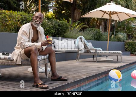 Homme afro-américain mature en chemise, short assis près de la piscine tenant cocktail jaune, espace de copie Banque D'Images