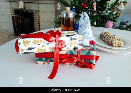 Deux cadeaux festifs enveloppés dans un motif d'arbre doré et du papier géométrique avec un arc rouge, empilés à côté d'une presse française en verre de thé et stollé devant un glo Banque D'Images