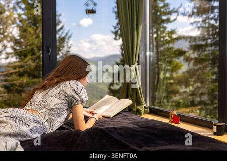 Jeune femme lisant par la fenêtre panoramique de montagne. Jeune femme est allongée sur un lit lisant un livre ouvert à côté d'une fenêtre panoramique, entourée par la lumière du soleil Banque D'Images