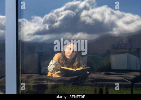 Jeune femme lisant derrière la fenêtre panoramique réfléchissante. Jeune femme lit un livre ouvert tout en se reposant sur un lit derrière une grande fenêtre réfléchissante, avec Banque D'Images