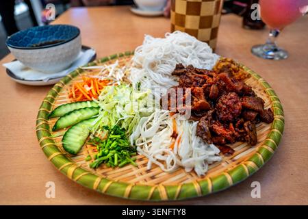 Repas vietnamien Bun cha (morceau de poitrine de porc grillé, légumes et nouilles de riz) sur une assiette décorative, bol avec sauce, limonade sur table. Banque D'Images