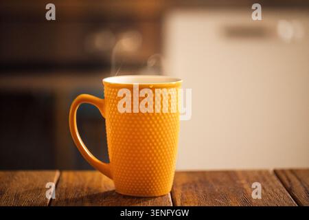 Tasse jaune avec un design texturé tient une boisson chaude sur une table en bois, avec des appareils de cuisine légèrement flous en arrière-plan Banque D'Images