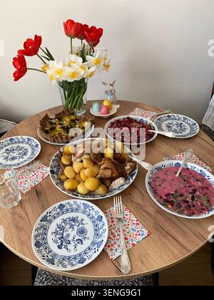 Une table est remplie de divers plats pour une célébration de Pâques. Il y a des salades, de la viande, des pommes de terre et des fleurs colorées disposées dans une salle à manger confortable Banque D'Images