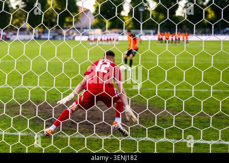 Regardez à travers le filet d'un gardien de but de football dans un kit rouge se préparant à défendre un penalty lors d'un match de football. Banque D'Images