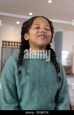 Enfant femelle debout et riant dans la cuisine à la maison portant un pull sarcelle et des liens de cheveux violets Banque D'Images