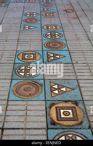 Un sentier géométrique en mosaïque de cercles, de carrés et de triangles dans des tons bleus et terreux est intégré dans une passerelle piétonne en carreaux de pierre. Banque D'Images