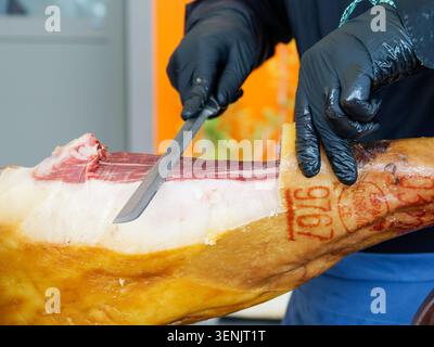 Gros plan d'un professionnel tranchant du jambon séché à partir d'une jambe de jambon à l'aide d'un couteau, typique de la charcuterie européenne, en France. Banque D'Images