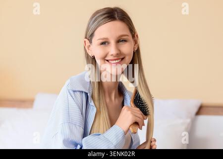 Jeune femme souriante brossant les longs cheveux blonds avec peigne en bois, routine de soins capillaires et traitement de beauté Banque D'Images