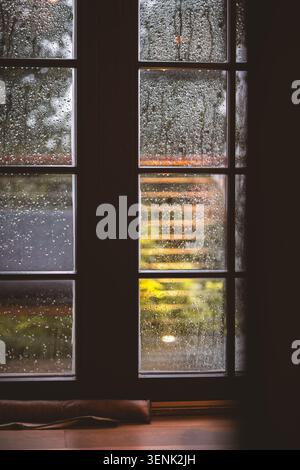 Vue rapprochée d'une fenêtre couverte de gouttes de pluie, avec un accent doux sur le paysage extérieur flou et des lumières chaudes créant une atmosphère chaleureuse Banque D'Images