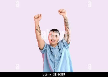 Jeune homme excité avec des tatouages levant les bras dans la célébration, exprimant la joie et le succès sur un fond clair Banque D'Images