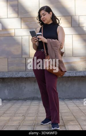 Femme adulte portant un haut noir, un pantalon marron, debout sur la plaza vérifiant le smartphone et tenant la tasse Banque D'Images