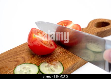 Gros plan d'un couteau de chef en métal tranchant coupant une tomate rouge mûre sur une planche de bois avec des tranches de concombre à proximité Banque D'Images