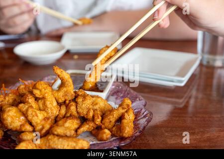 Gros plan d'action détaillé capturant la main d'une personne à l'aide de baguettes de bois pour tremper un morceau de poulet frit doré et croustillant (tempura) dans un smal Banque D'Images