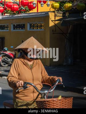 Femme vietnamienne âgée avec un chapeau conique à vélo dans une rue ensoleillée Banque D'Images