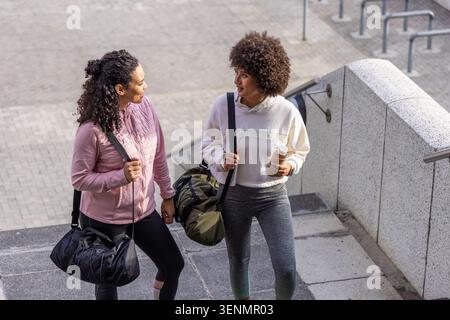 Deux amies en vêtements d'entraînement marchant dans l'escalier de plaza transportant des sacs marins et une bouteille Banque D'Images