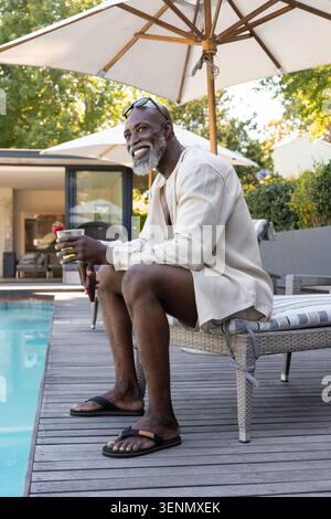 Homme africain senior assis sur une chaise longue tissée sur la terrasse en bois de la piscine, tenant un cocktail garni Banque D'Images