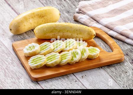 Concombres jaunes fraîchement tranchés disposés sur une planche à découper en bambou à côté de légumes entiers. Fond en bois rustique avec serviette de cuisine rayée ajoute Banque D'Images