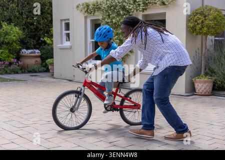 Homme afro-américain Steadying Boy portant un casque bleu sur un vélo rouge sur l'allée, en chemise à carreaux Banque D'Images