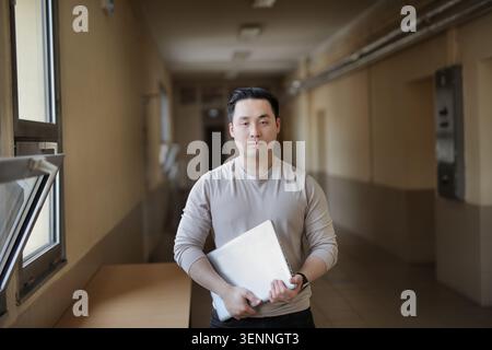 Étudiant asiatique souriant tenant un ordinateur portable dans le couloir de l'université Banque D'Images