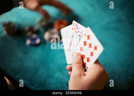 La main affiche des cartes à jouer avec des diamants rouges tandis que les jetons de poker sont dispersés sur une table en feutre vert, créant une atmosphère de jeu décontractée Banque D'Images