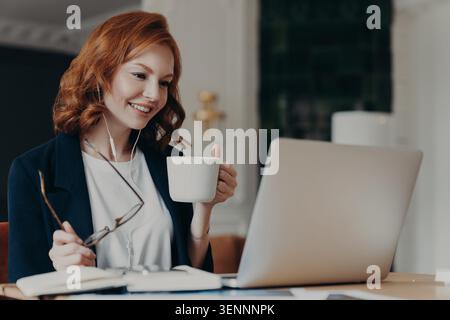 Femme d'affaires aux cheveux rouges boit du café tout en travaillant sur un ordinateur portable à un bureau Banque D'Images