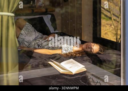Femme se reposant à côté du livre ouvert dans la cabine ensoleillée. Jeune femme dans une robe à motifs repose les yeux fermés sur un lit sombre à l'intérieur d'une cabane en bois Banque D'Images