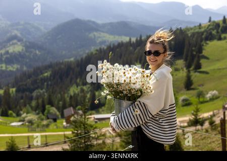 Femme souriante tenant des Marguerites dans le paysage de montagne verte. Jeune femme souriante en lunettes de soleil et un pull rayé tient un seau en métal rempli de Banque D'Images