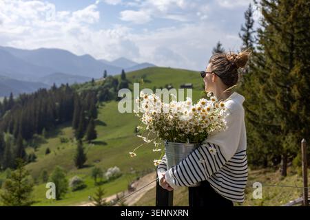 Femme tenant des marguerites surplombant Green Mountain Hills. Jeune femme en lunettes de soleil et un pull rayé tient un seau en métal débordant de blanc Banque D'Images