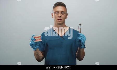 Homme dentiste dans les gommages bleus tient miroir dentaire et modèle de dent avec les mains gantées dans le studio ; calme professionnel. Banque D'Images