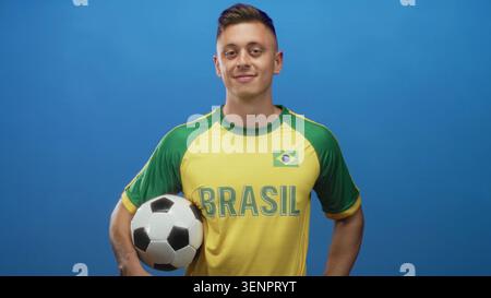 Homme portant le maillot brésilien tient le ballon de football avec le bras et le sourire, pose avec confiance en studio avec fond bleu ; fierté camaraderie. Banque D'Images